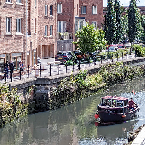 Pedestrians outside BottleWorks while a boat sails past on the Ouseburn river in Newcastle upon Tyne